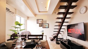 A modern living room with a large flat-screen TV displaying 'NETFLIX', a dining area with a table set for two, and a staircase with a wooden railing. - Fort Victoria BGC Netflix 25M WiFi (Taguig)