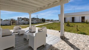 Een terras buiten met witte wicker meubels en een stenen vloer, onder een witte pergola, met een modern gebouw op de achtergrond.