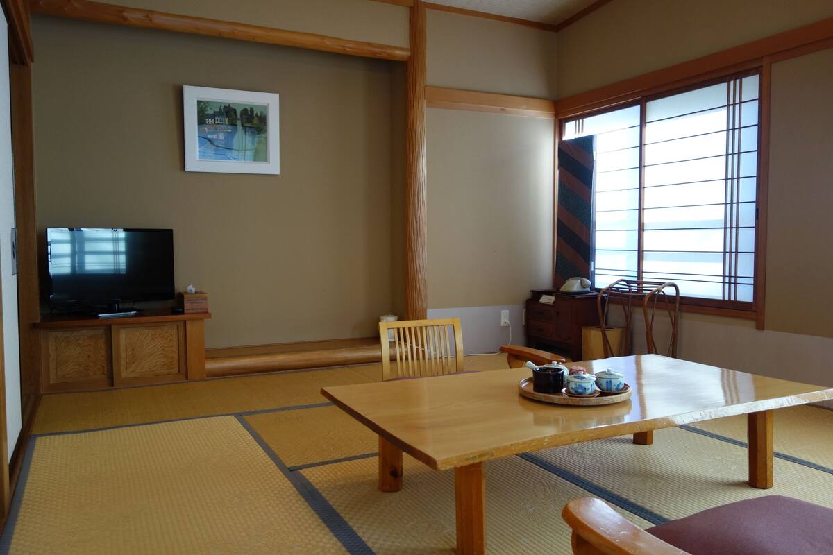 Una habitación japonesa tradicional con suelo de tatami, una mesa de madera con bandeja de té, un televisor sobre un mueble bajo y una puerta corredera con vistas.
