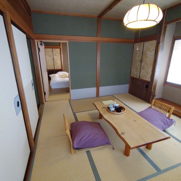 Una habitación tradicional japonesa con suelo de tatami, una mesa de madera baja con una bandeja de utensilios de té y una cama con un cojín morado.