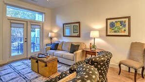 A living room with a sofa, armchairs, a coffee table, and a side table with a lamp. There is a door leading to an outdoor patio area with chairs.