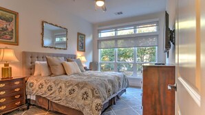 A bedroom with a large bed, a dresser, a mirror, and a window with a view of greenery.