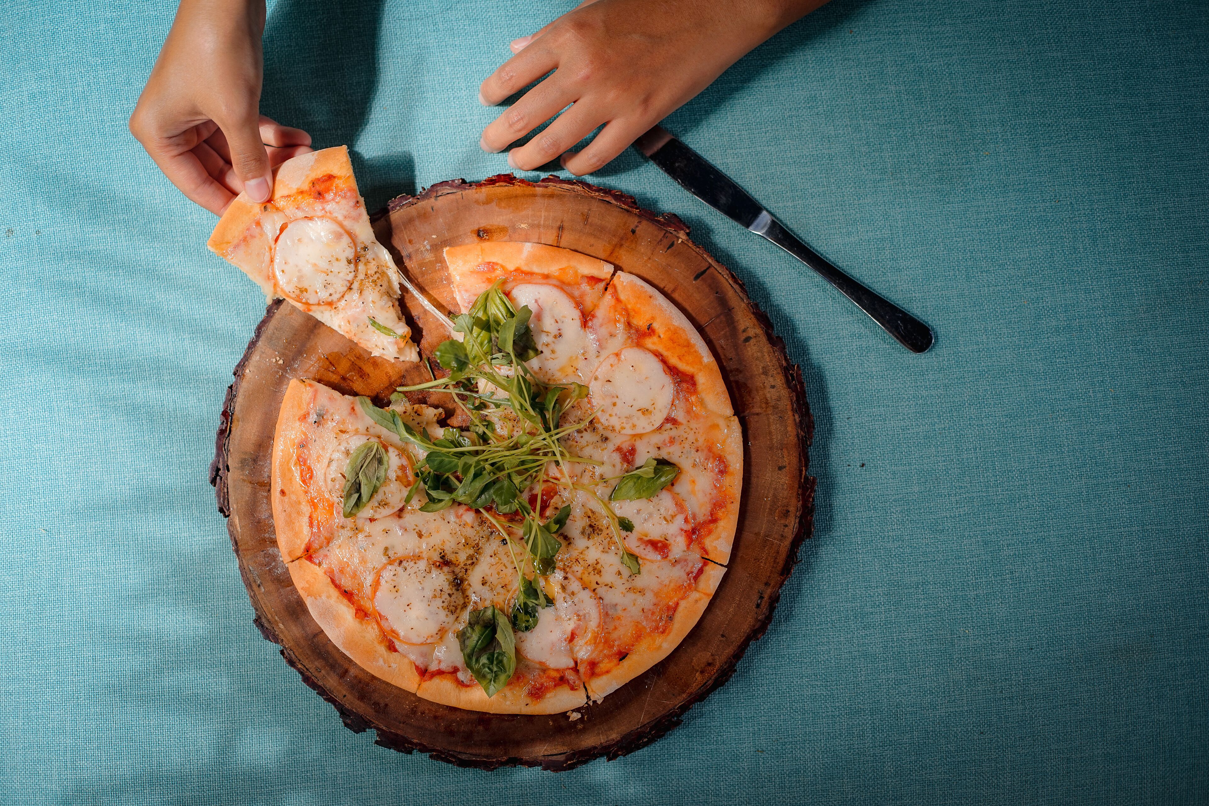 A pizza on a wooden board with a knife and a hand picking up a slice.