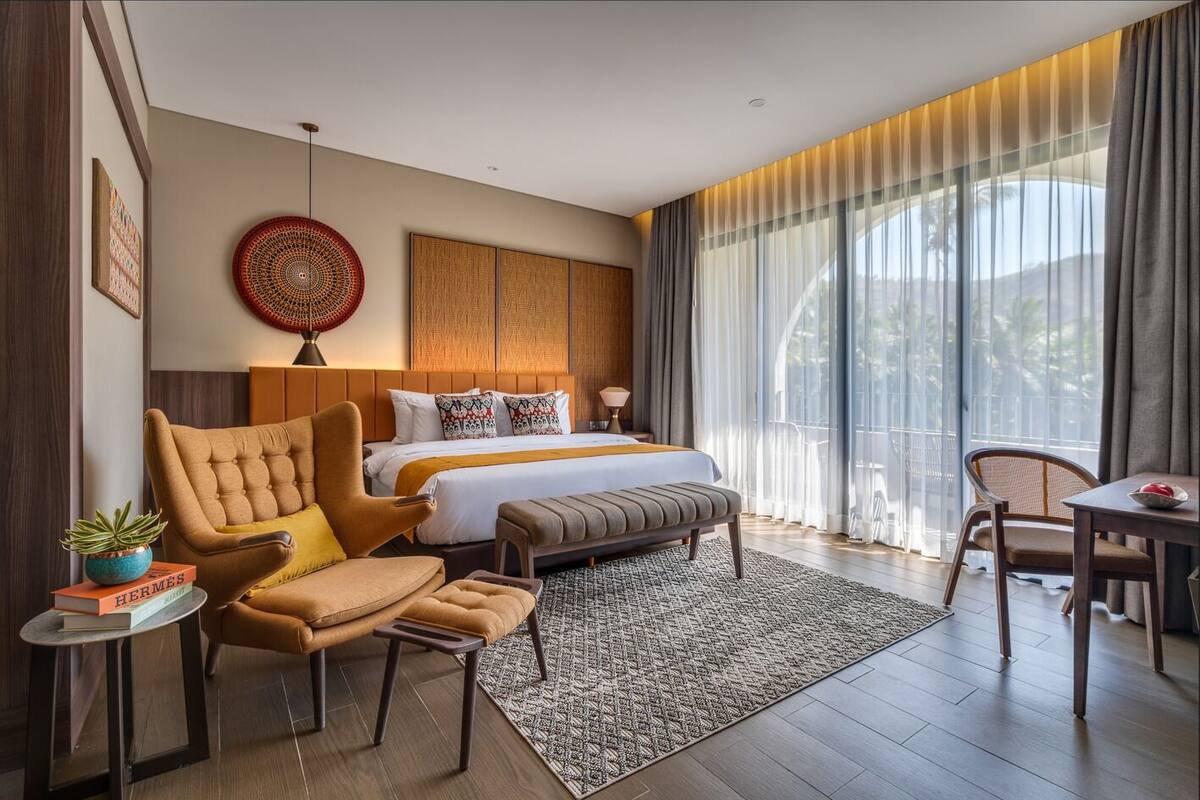 Ein modernes Hotelzimmer mit einem großen Bett, einer Sitzecke mit Stuhl und Tisch sowie Blick durch große Fenster.