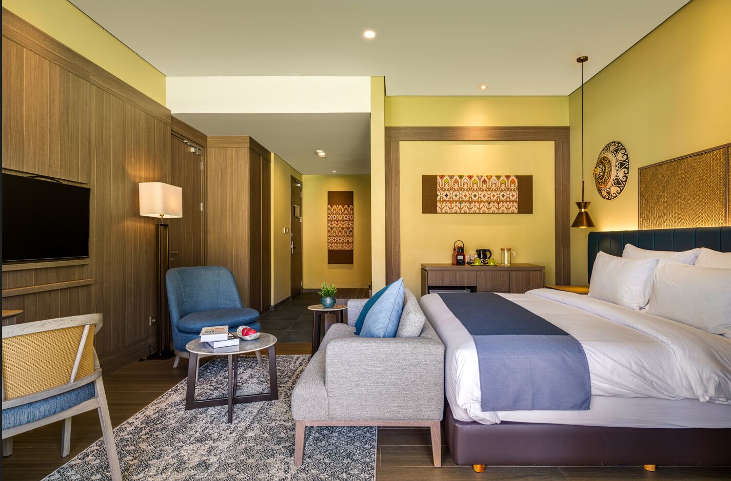 Habitación de hotel con cama, sofá, sillón, televisión y una mesita con lámpara y planta.
