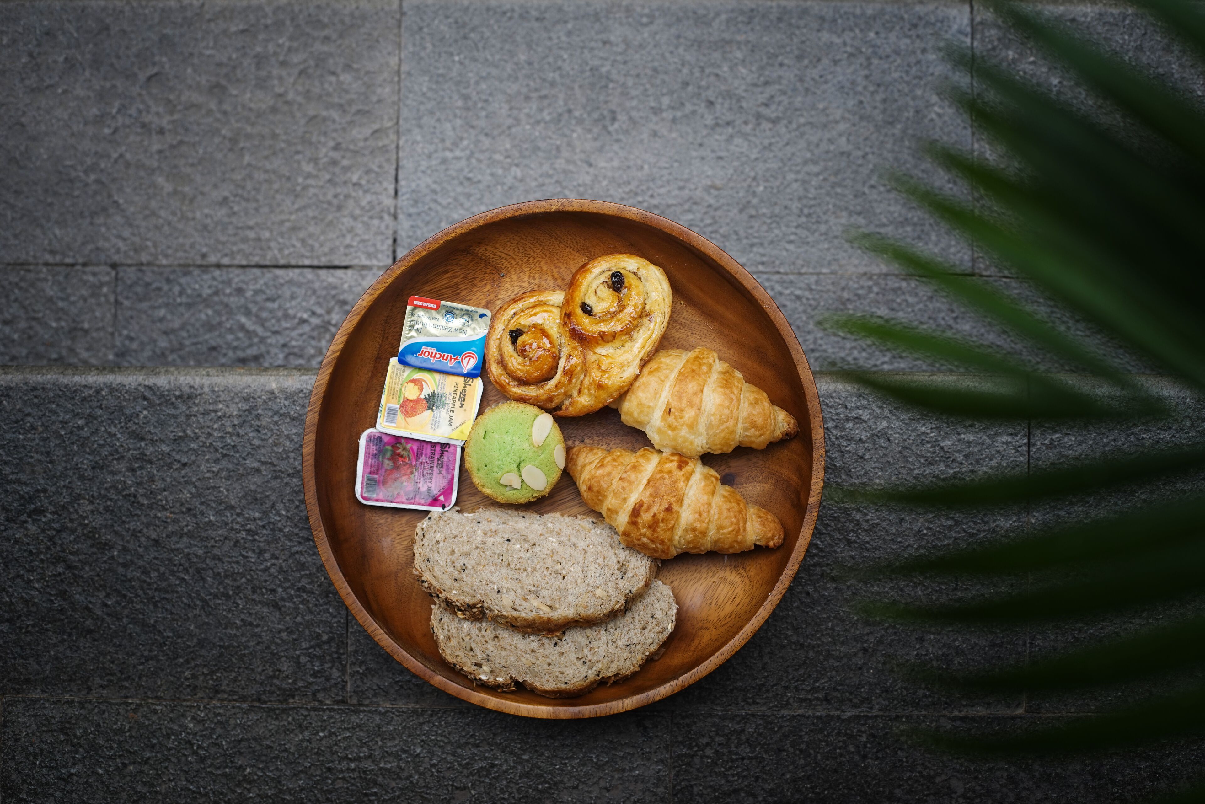 Ein hölzernes Tablett mit Croissants, grüner Creme und einem kleinen Marmeladenglas auf einer strukturierten Steinoberfläche.