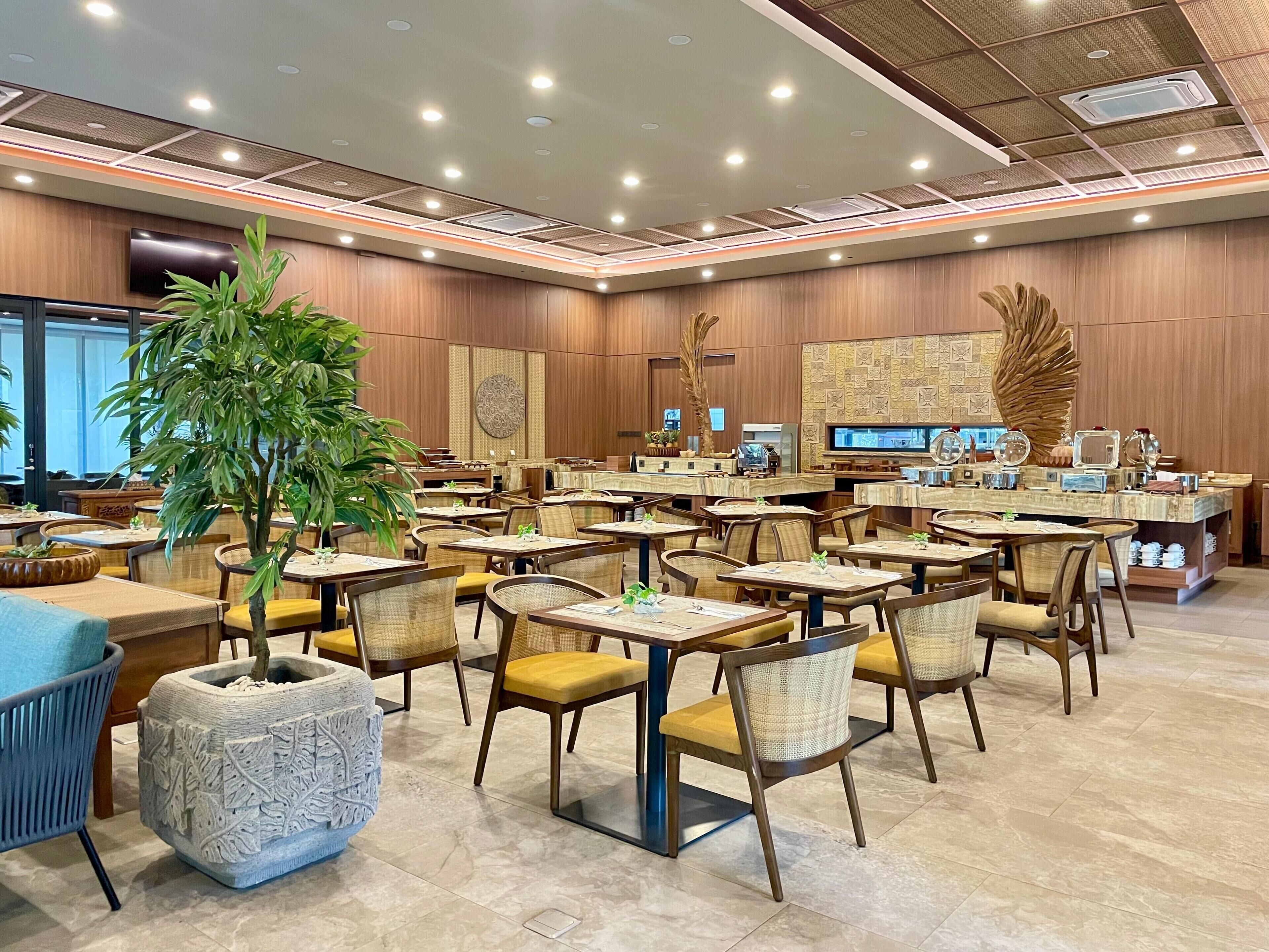 A spacious restaurant with multiple tables and chairs, a large decorative plant, and a modern ceiling design.