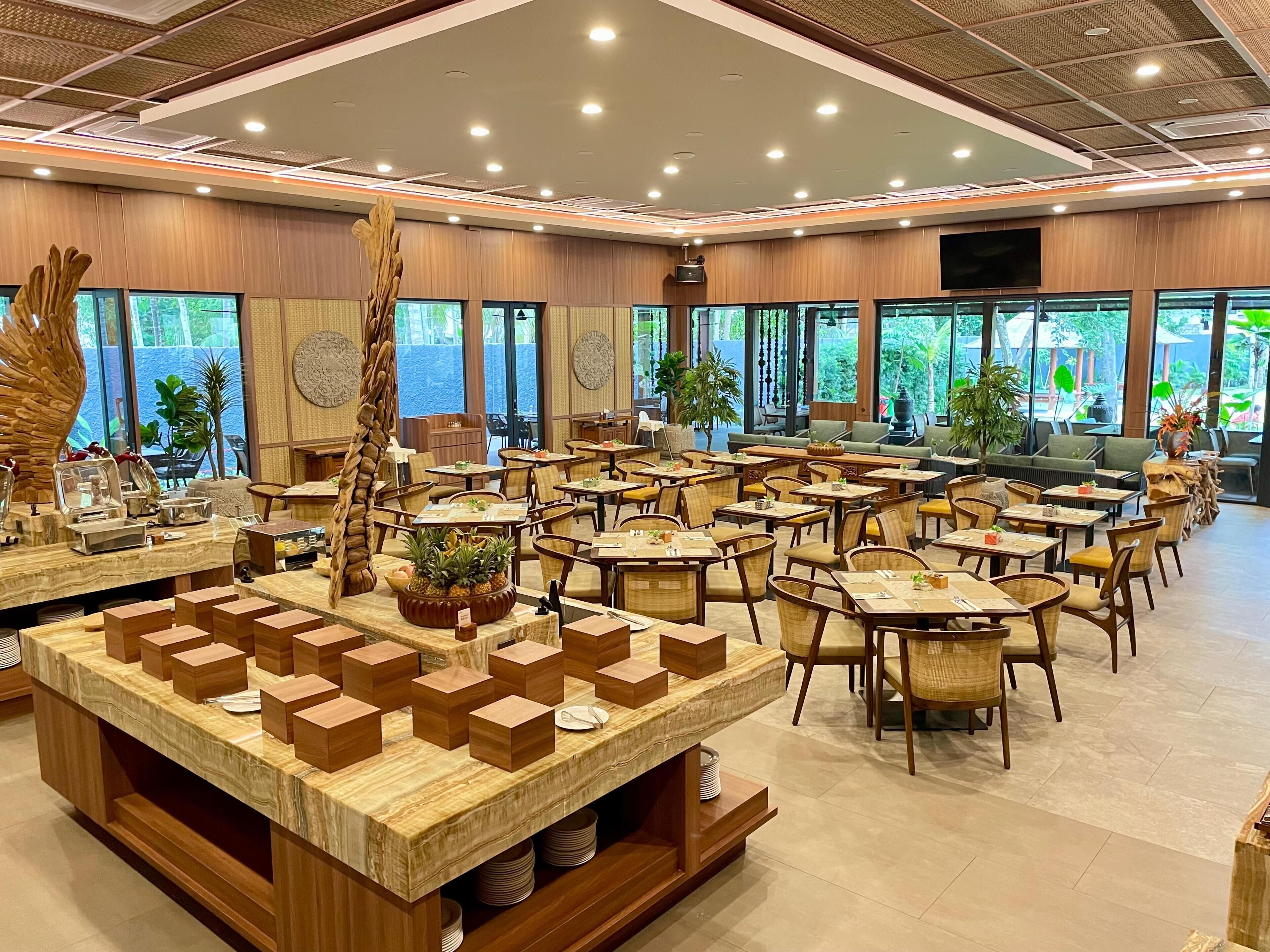 A spacious restaurant with multiple tables and chairs, a central island with a fruit bowl, and large windows offering a view of the outdoors.