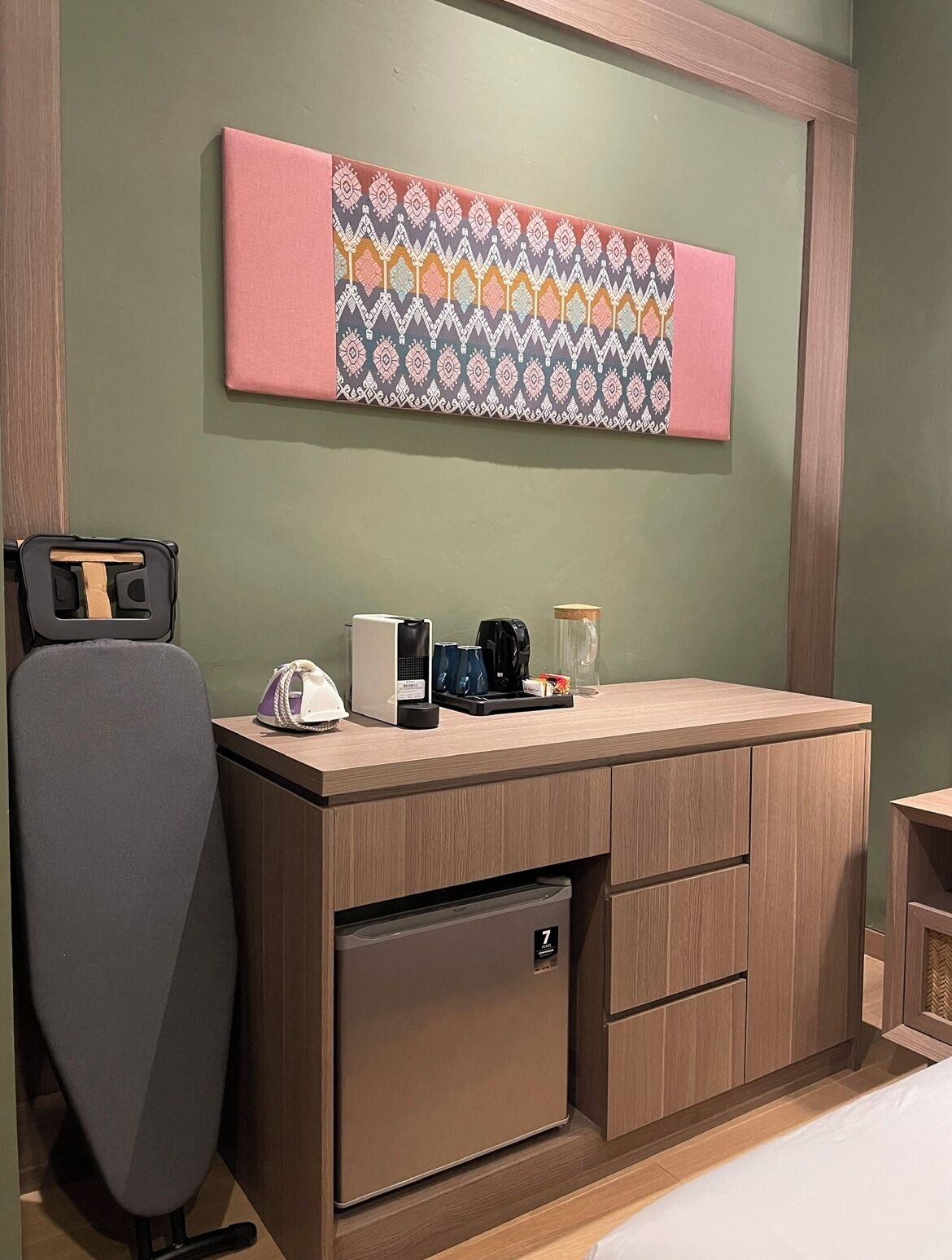 A compact hotel room with a wooden desk, a small refrigerator, a coffee maker, and a decorative wall hanging.