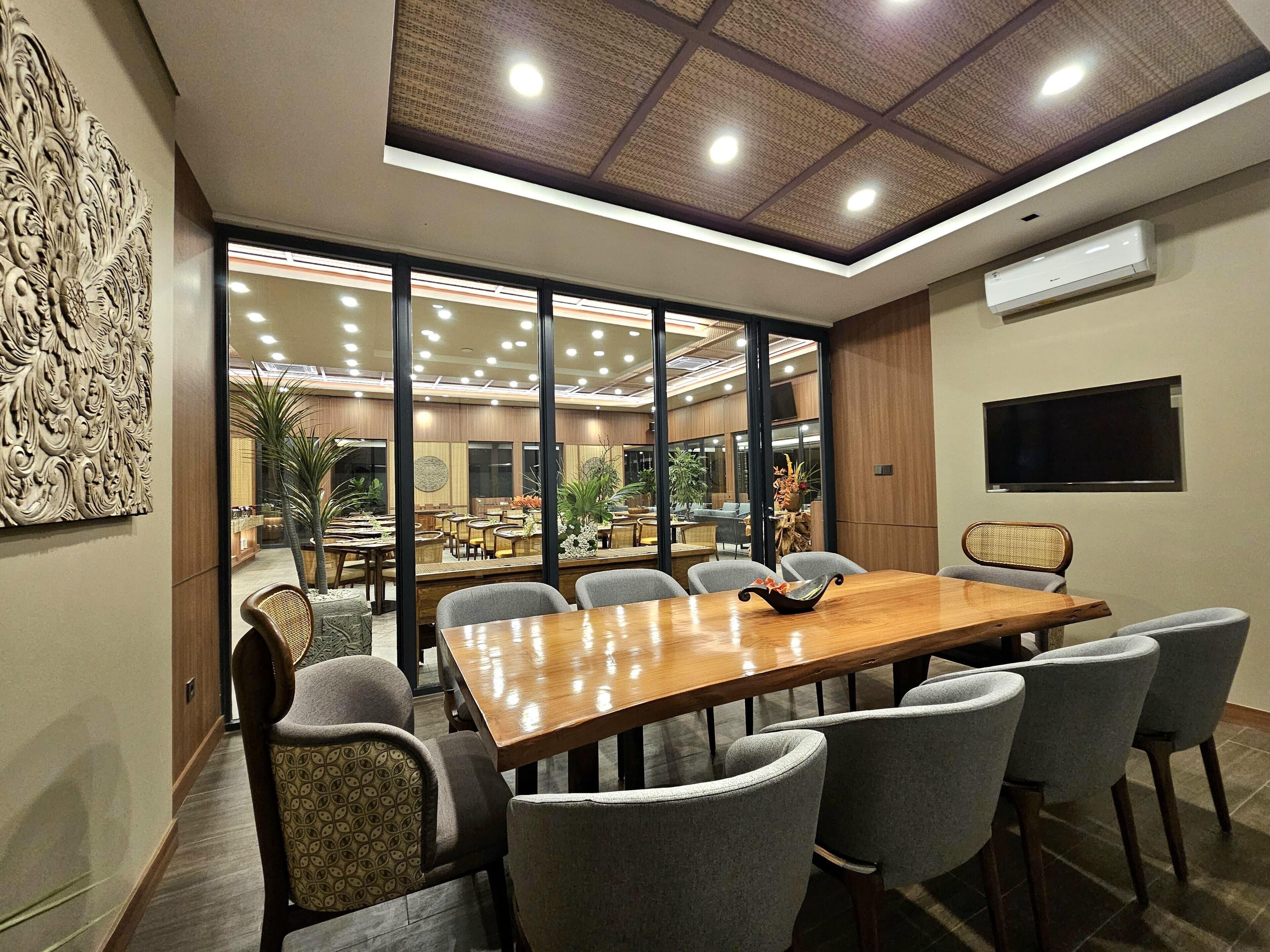 Un salón de conferencias con una gran mesa de madera, sillas tapizadas en gris, un techo con iluminación empotrada y un amplio ventanal con vistas al comedor.