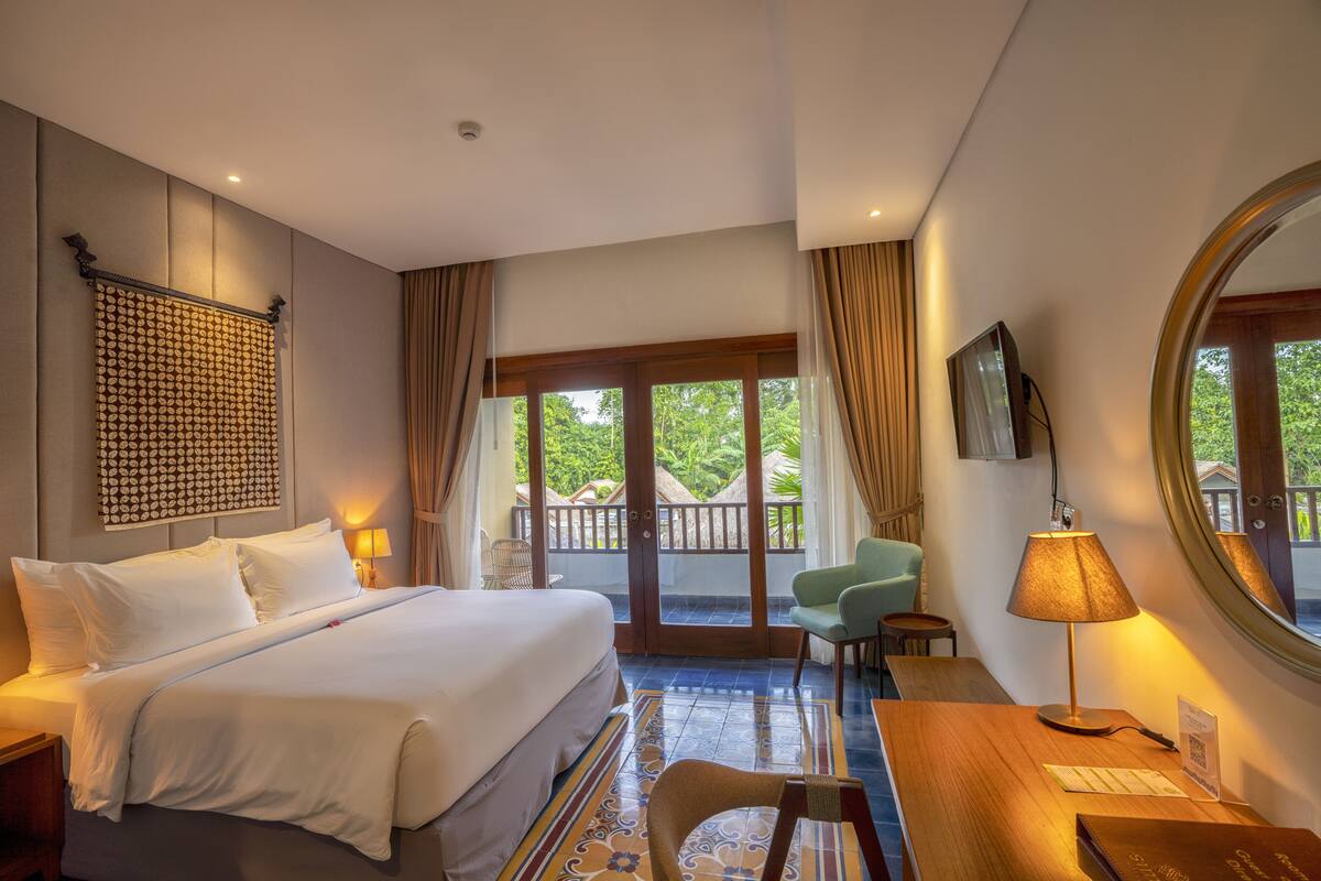 Ein Hotelzimmer mit einem großen Bett, einem Schreibtisch, einem Sessel, einem Fernseher und einem Balkon mit Blick auf Grünflächen.
