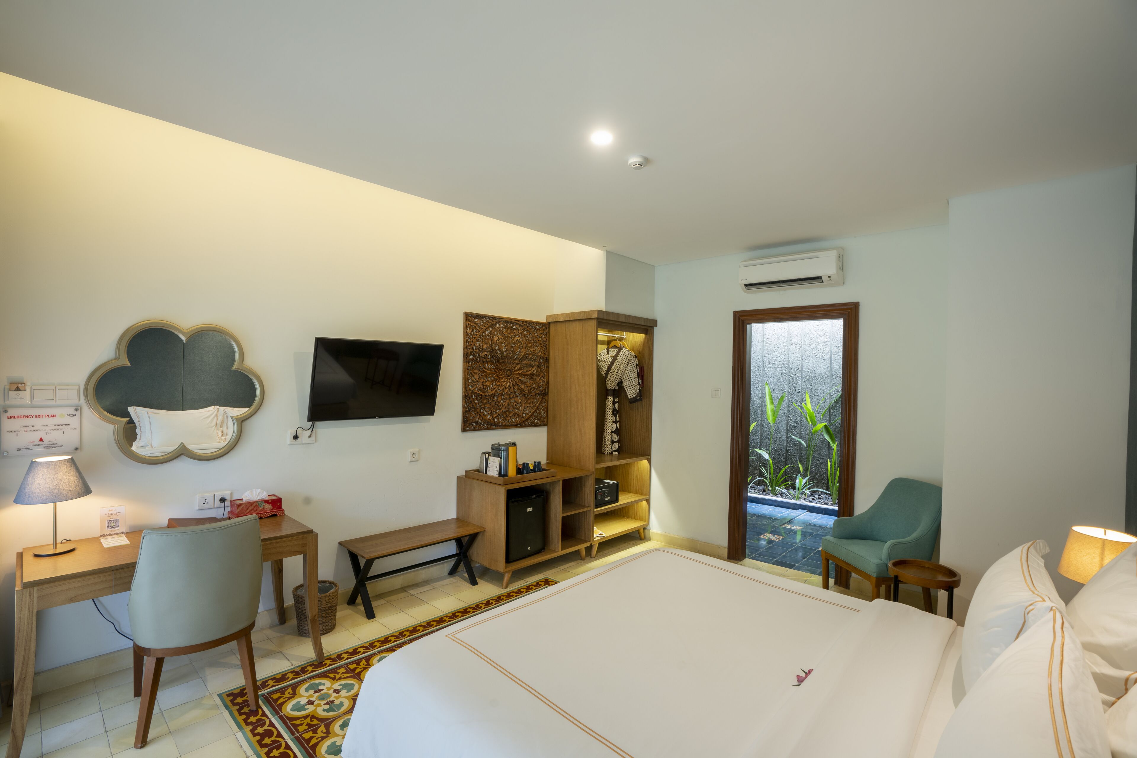 Quarto de hotel com cama, escrivaninha, cadeira e um pequeno espaço de estar com vista para plantas lá fora.