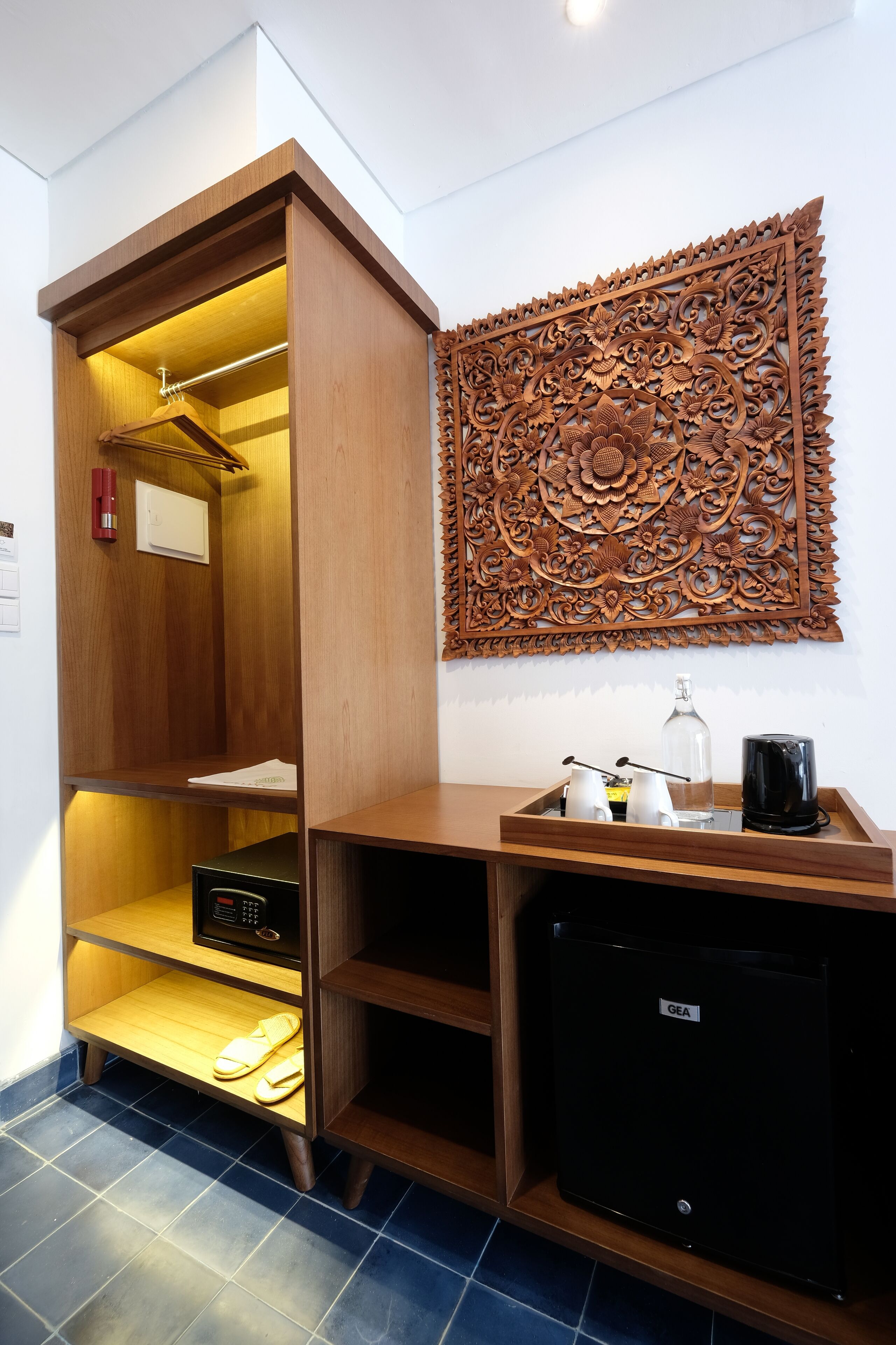 A compact hotel room with a wooden wardrobe, a small black refrigerator, a decorative wall panel, and a bedside table with a bottle and glasses.