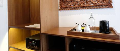 A compact hotel room with a wooden wardrobe, a small black refrigerator, a decorative wall panel, and a bedside table with a bottle and glasses.