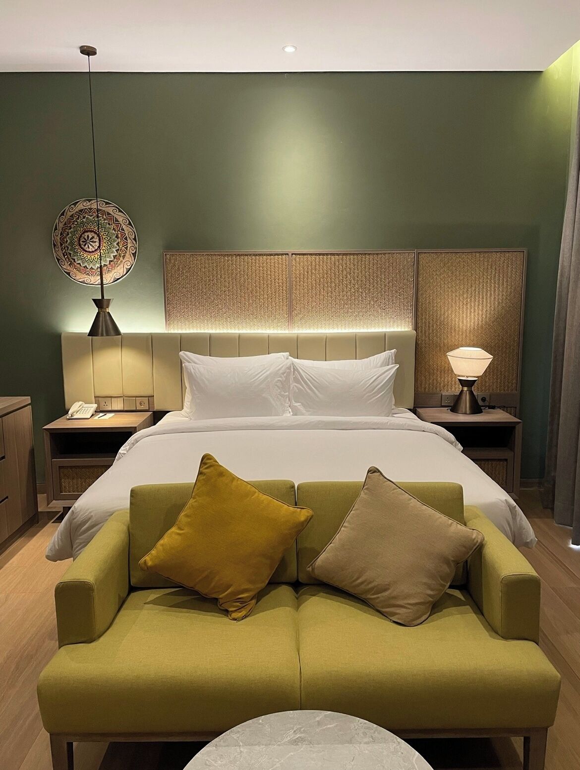 A hotel room with a bed, a green sofa, a bedside table with a lamp, and a decorative wall panel.