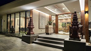 Una entrada de edificio moderno con fachada de vidrio, columnas de madera y una escalera central que lleva a un comedor.