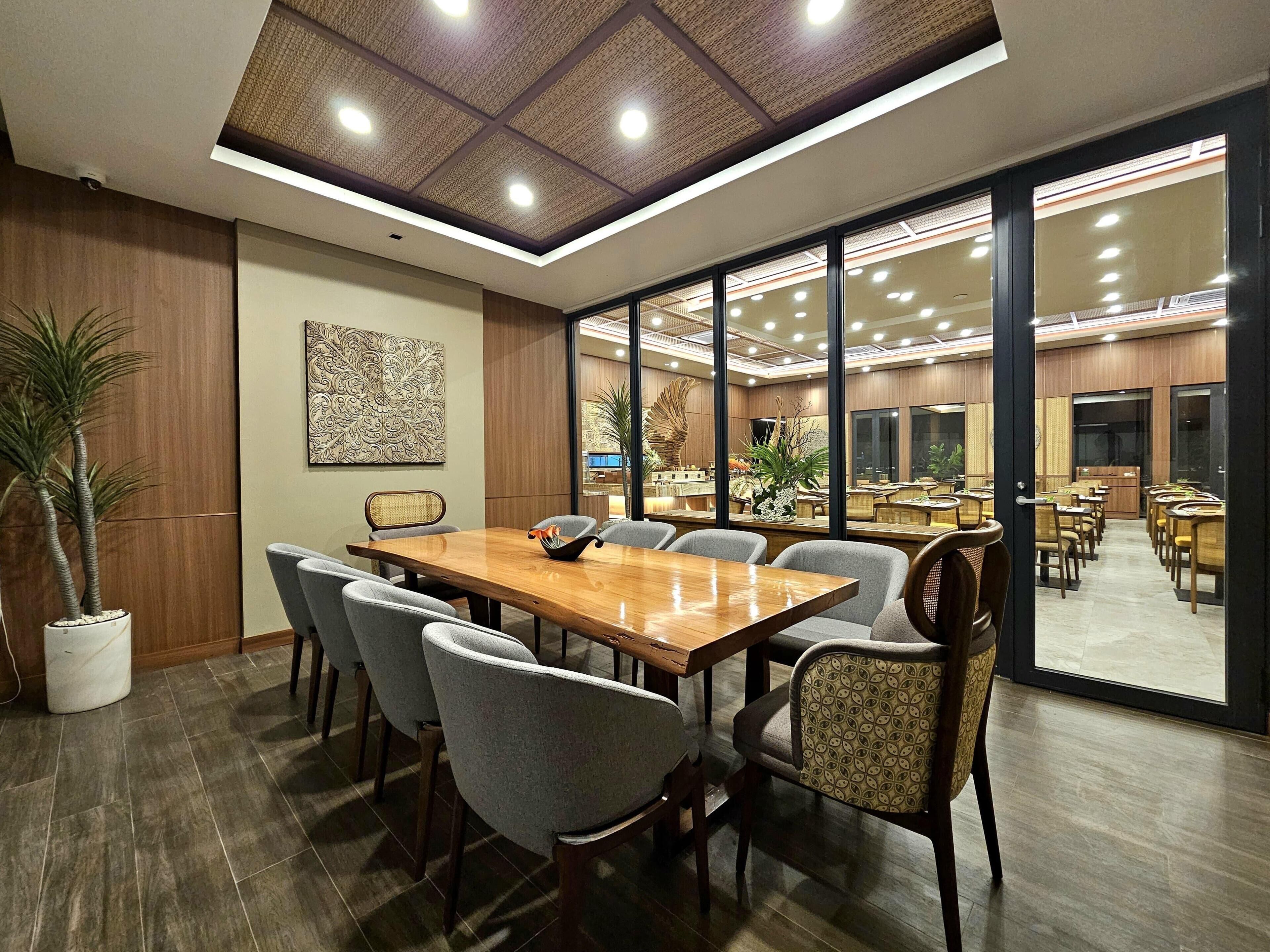 A conference room with a wooden table, grey chairs, a potted plant, and a decorative wall piece.