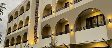 Ett hotell med flera våningar, en symmetrisk fasad, flera balkonger och ett välskött trädgårdsområde med gångvägar och små träd.