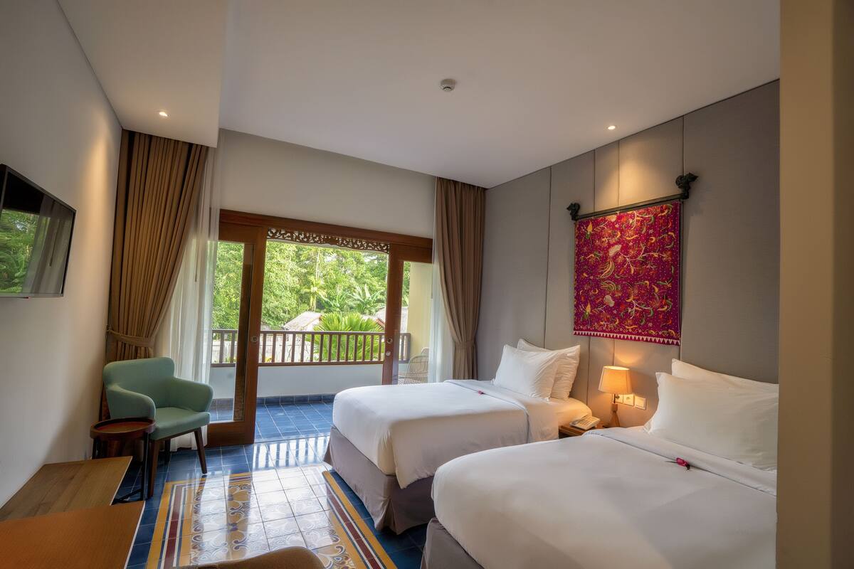 Ein Hotelzimmer mit zwei Betten, einem Sessel, einem kleinen Tisch, einem Balkon mit Vorhängen und einem Wandbehang.