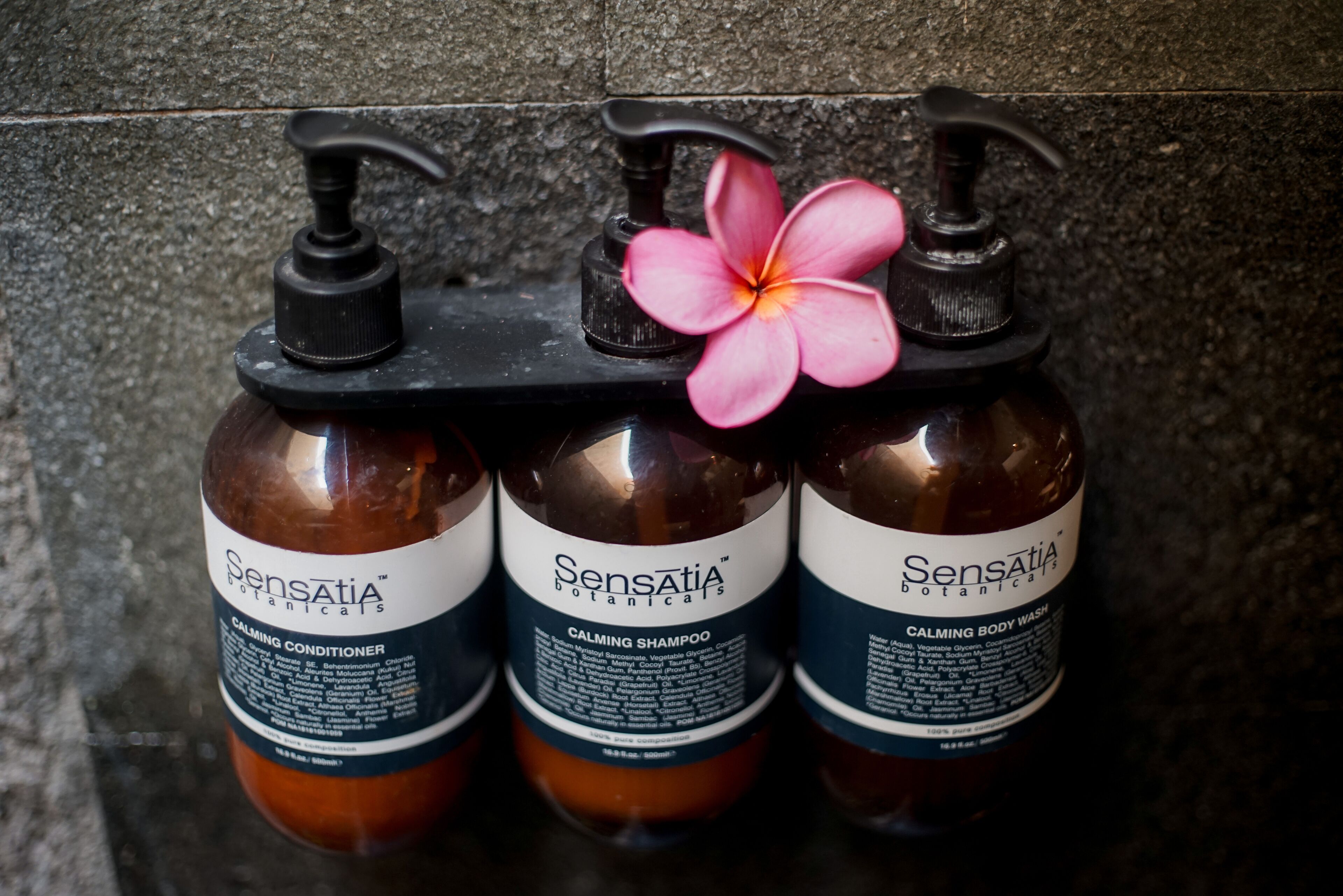 Three bottles of Sensatia Botanicals calming products on a black pump dispenser against a textured wall.