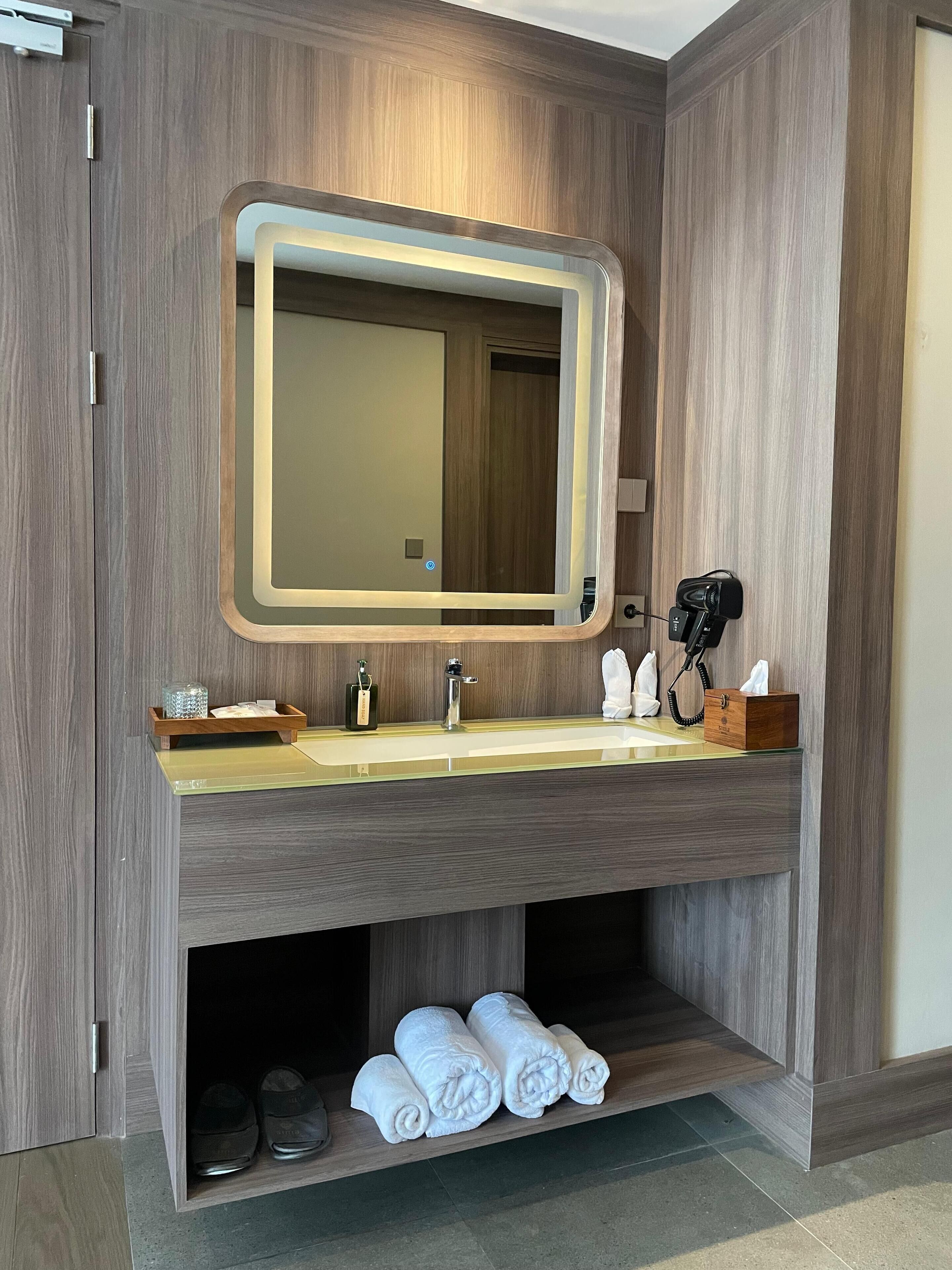 Une salle de bain moderne avec un meuble-lavabo en bois, un grand miroir, un sèche-cheveux et des serviettes soigneusement pliées.