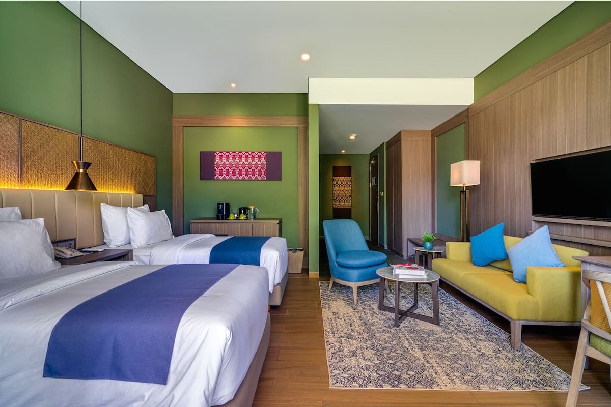 Ein Hotelzimmer mit zwei Betten, einem Schlafsofa, einem Sessel, einem Couchtisch, einem Fernseher und einer Wand mit einem Wandbild.