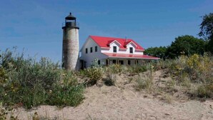A lighthouse with a red roof and a white building with a similar red roof, surrounded by dry vegetation and a clear sky. - Charity Island Lightkeepers Home Big Charity Island (Tawas)