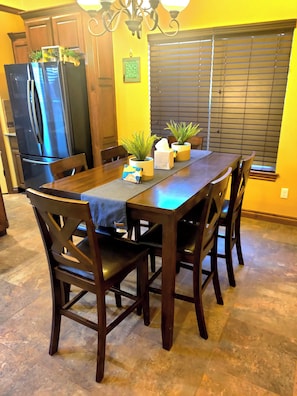 Un comedor con mesa de madera, sillas, candelabro, refrigerador y ventana con persianas.