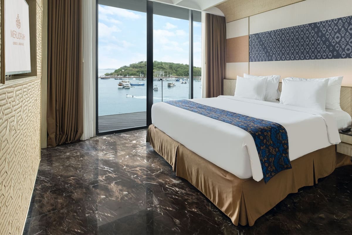 A hotel room with a large bed, a view of boats and greenery, a reception desk, and a marble floor.