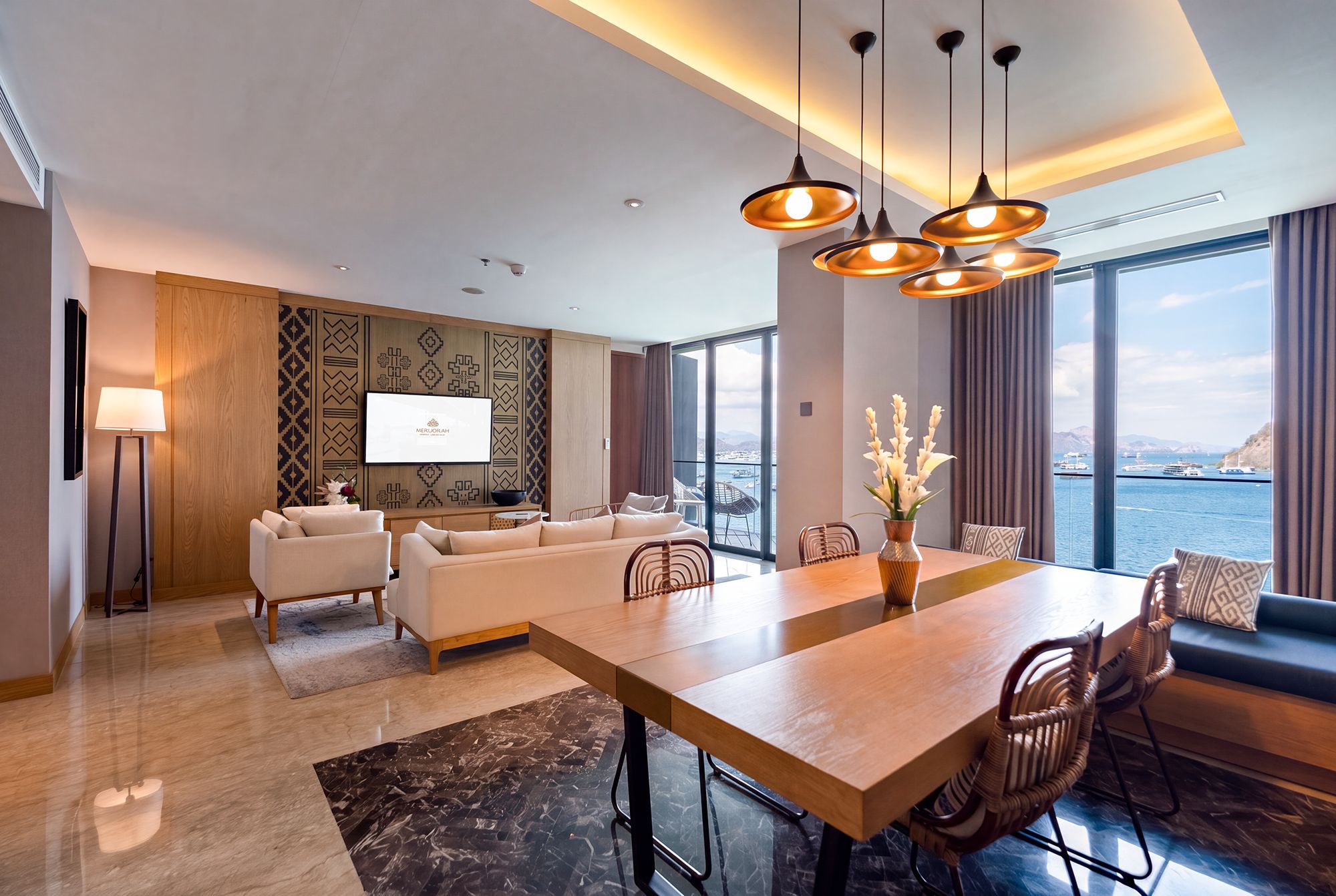 A modern hotel room with a large dining table, comfortable seating, a flat-screen TV, and a view of the sea.