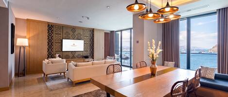 A modern hotel room with a large dining table, comfortable seating, a flat-screen TV, and a view of the sea.