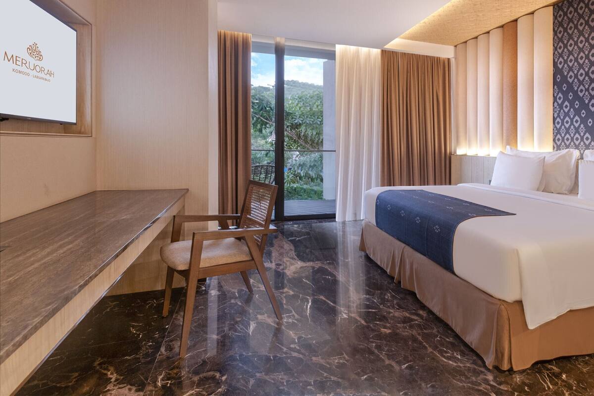 A hotel room with a bed, a desk, and a chair. There is a large window with curtains, and a wall-mounted TV.