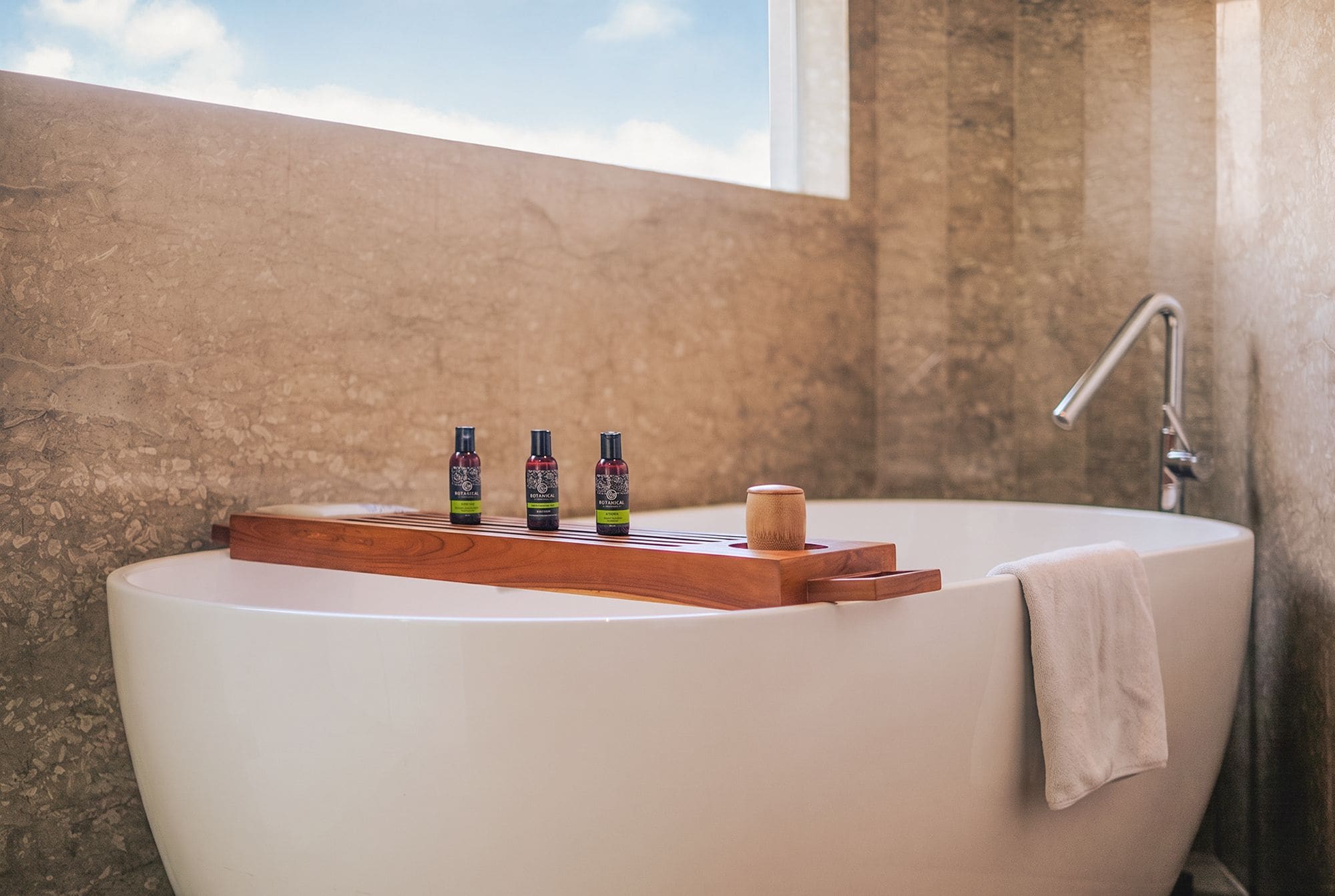 A bathtub with a wooden tray holding three bottles, a towel, and a window in the background.