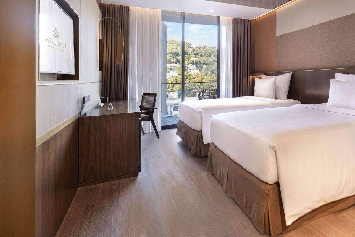 A hotel room with two beds, a desk, a chair, and a large window offering a view of the outdoors.