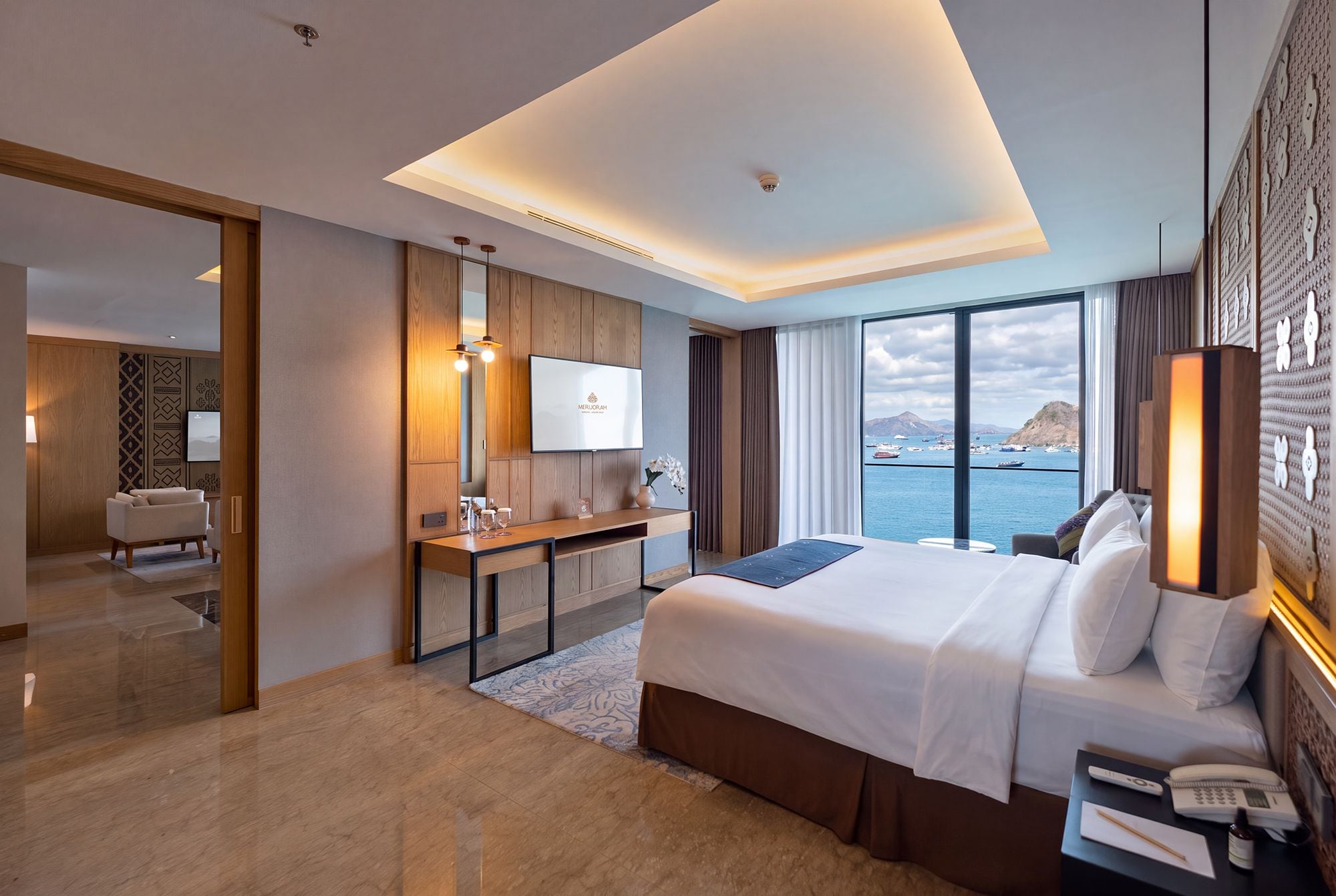A modern hotel room with a large bed, a desk, a television, and a view of the sea.