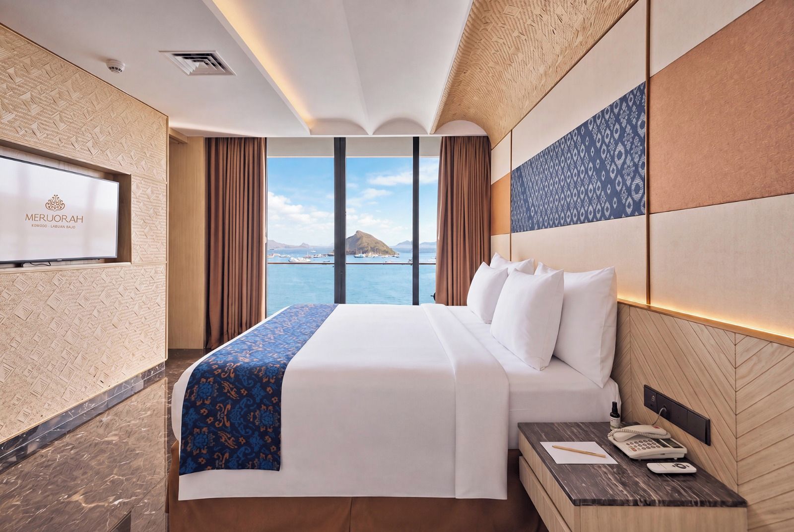 A hotel room with a bed, a desk with a phone, a TV, and a view of the sea.