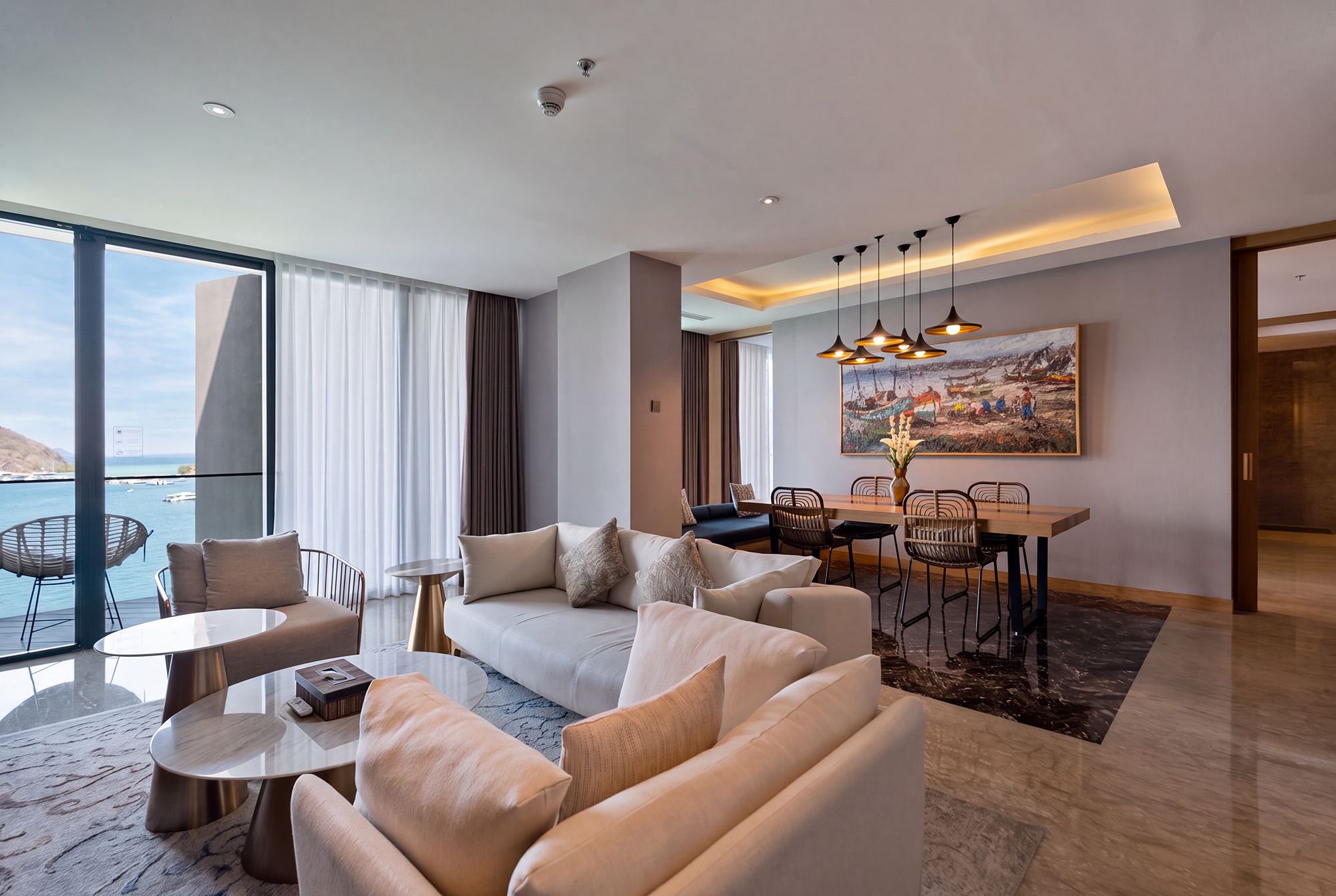A modern hotel room with a living area, dining space, and a large window offering a view of the sea.