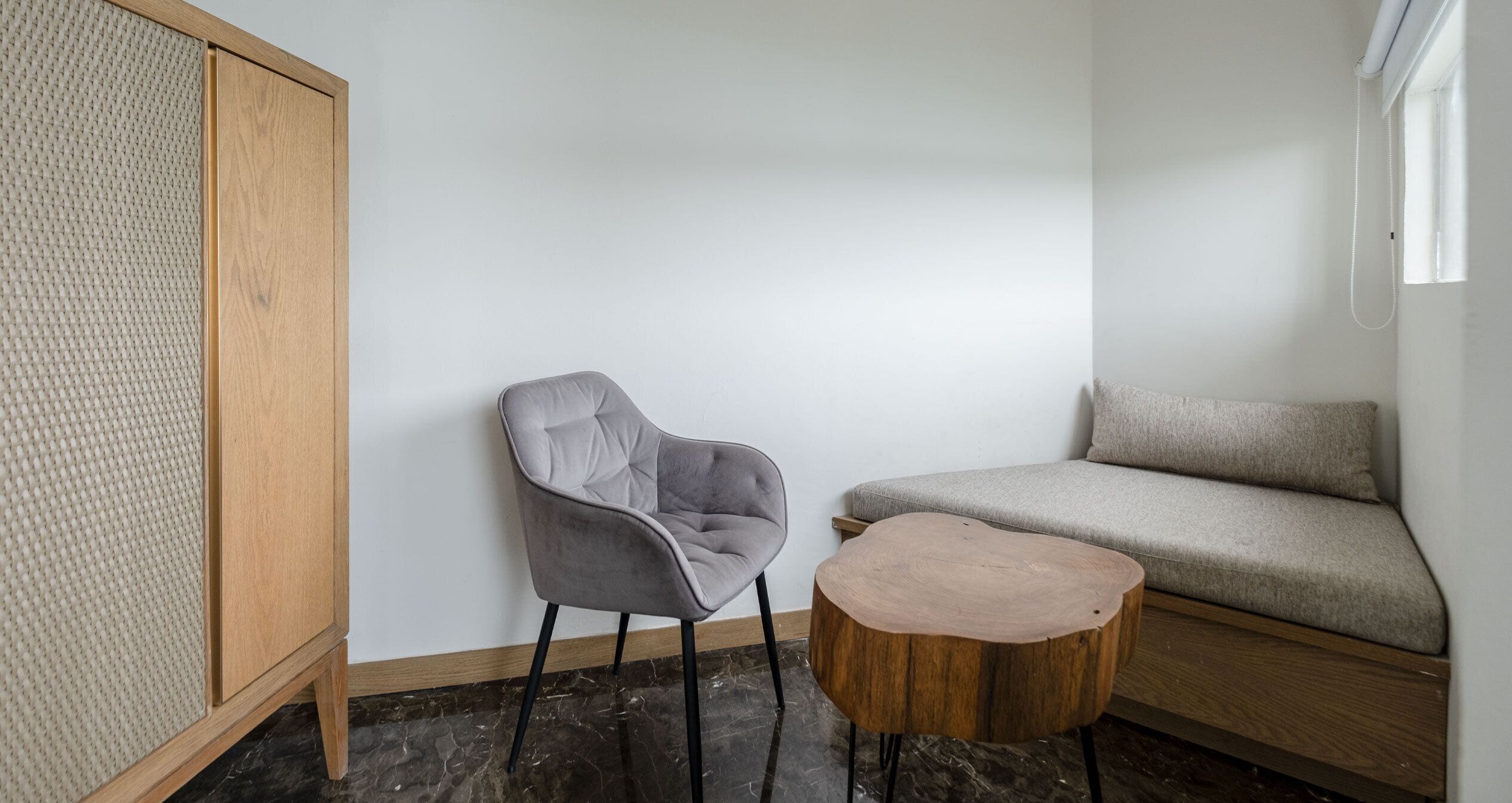 Una habitación con una silla tapizada de gris, una mesa redonda de madera, un sofá y un armario de madera.