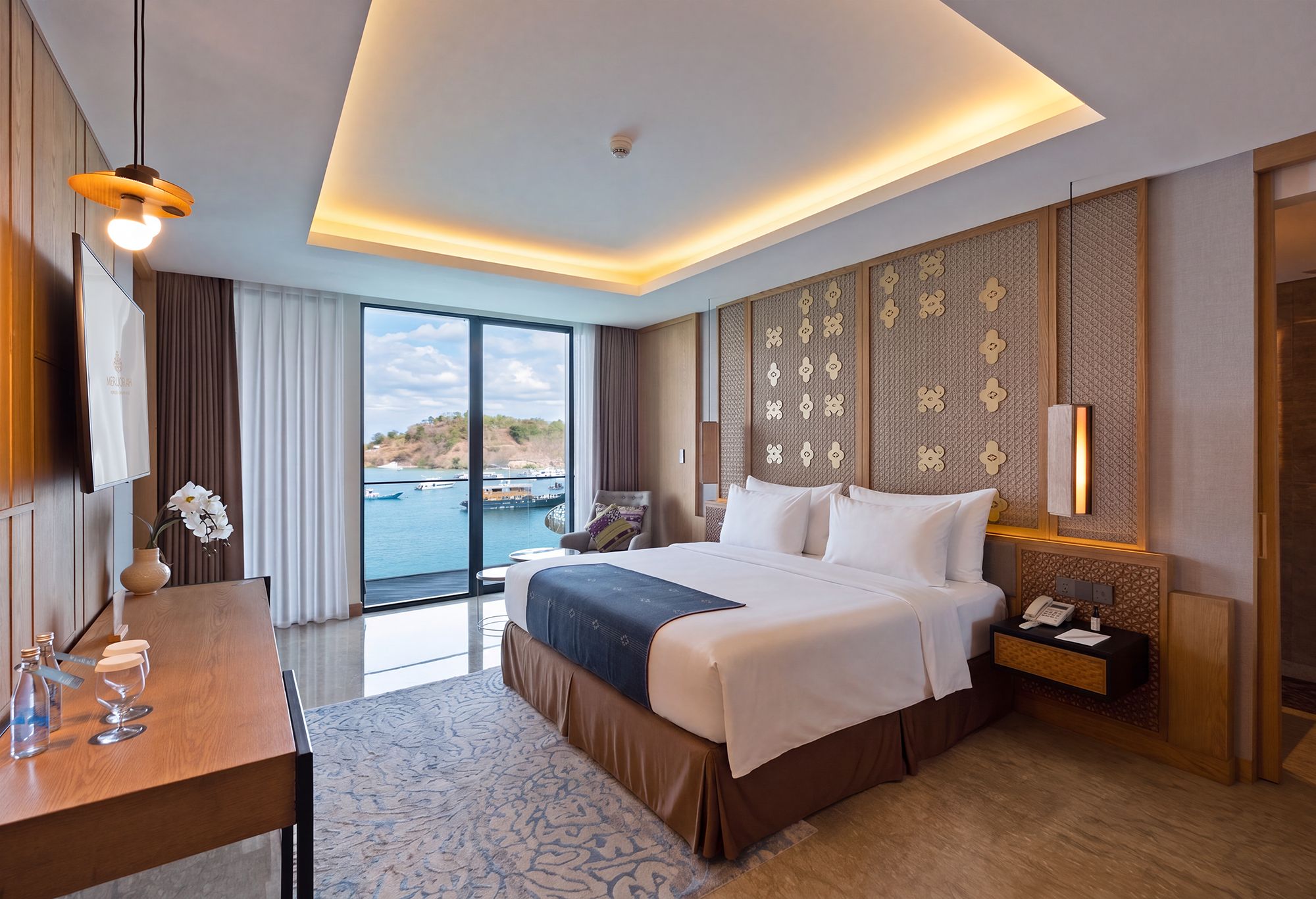A hotel room with a large bed, a desk with a vase of flowers, a nightstand, and a view of the sea.