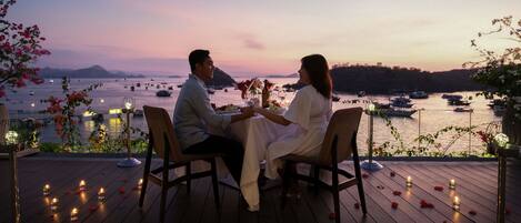 A couple seated at a table with a view of boats on a body of water, surrounded by flowers and candles.