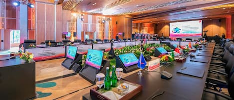 Un salón de conferencias con varias mesas, sillas y una pantalla grande mostrando una presentación.