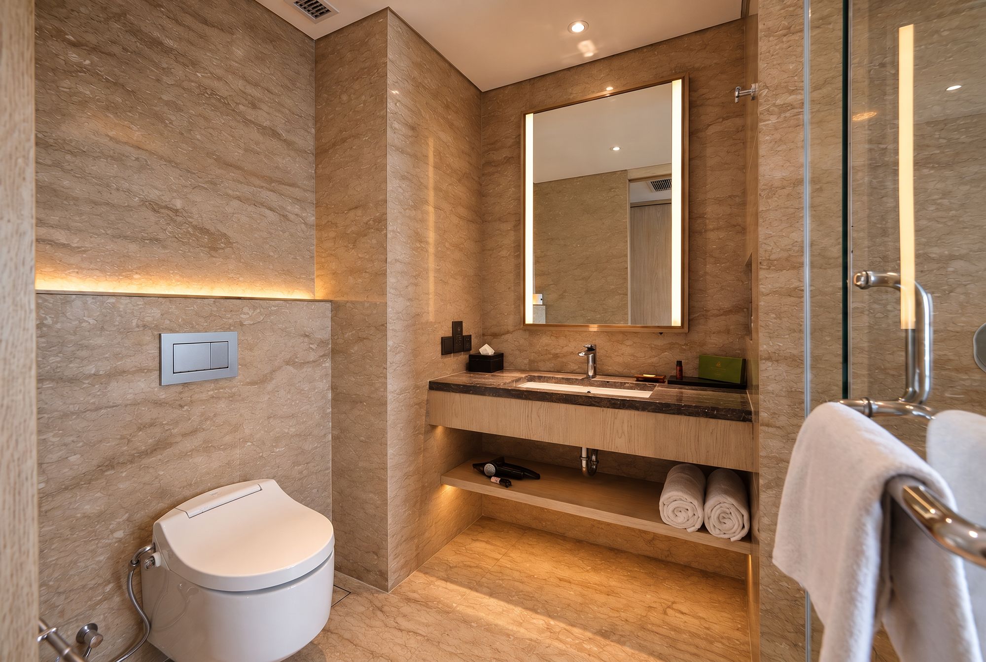 Un baño moderno con pared de mármol, un espejo grande, un lavamanos, un inodoro y una cabina de ducha de vidrio.
