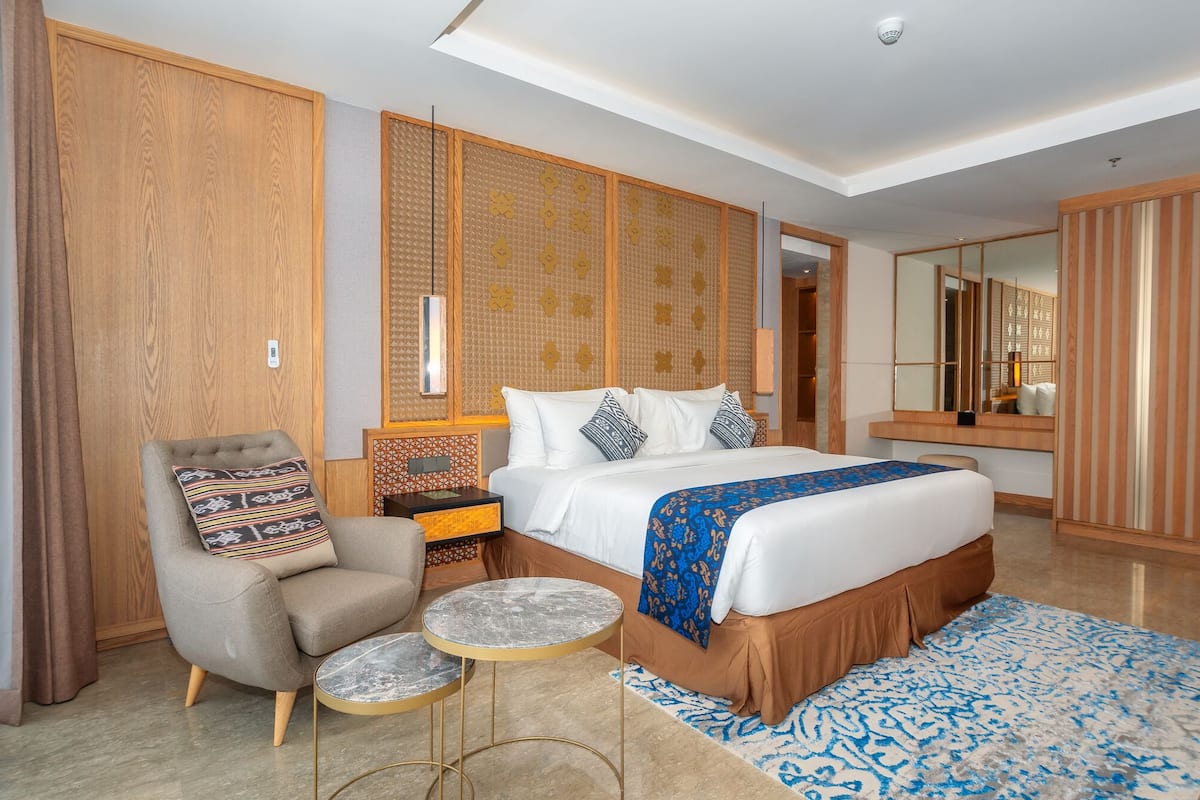 A modern hotel room with a large bed, a seating area with a chair and a small table, a bedside table with a lamp, and a large mirror.