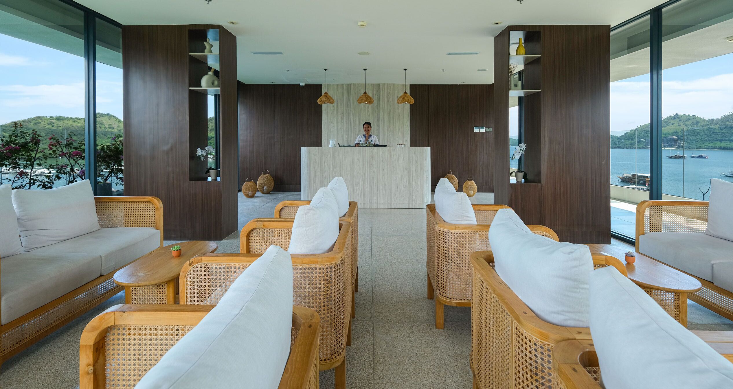 A modern reception area with wicker furniture, a wooden reception desk, and large windows offering a view of the sea and hills.