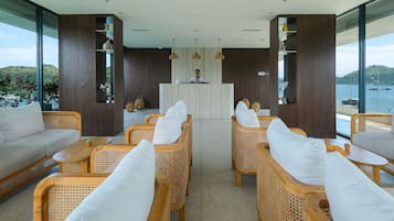A modern reception area with wicker furniture, a wooden reception desk, and large windows offering a view of the sea and hills.