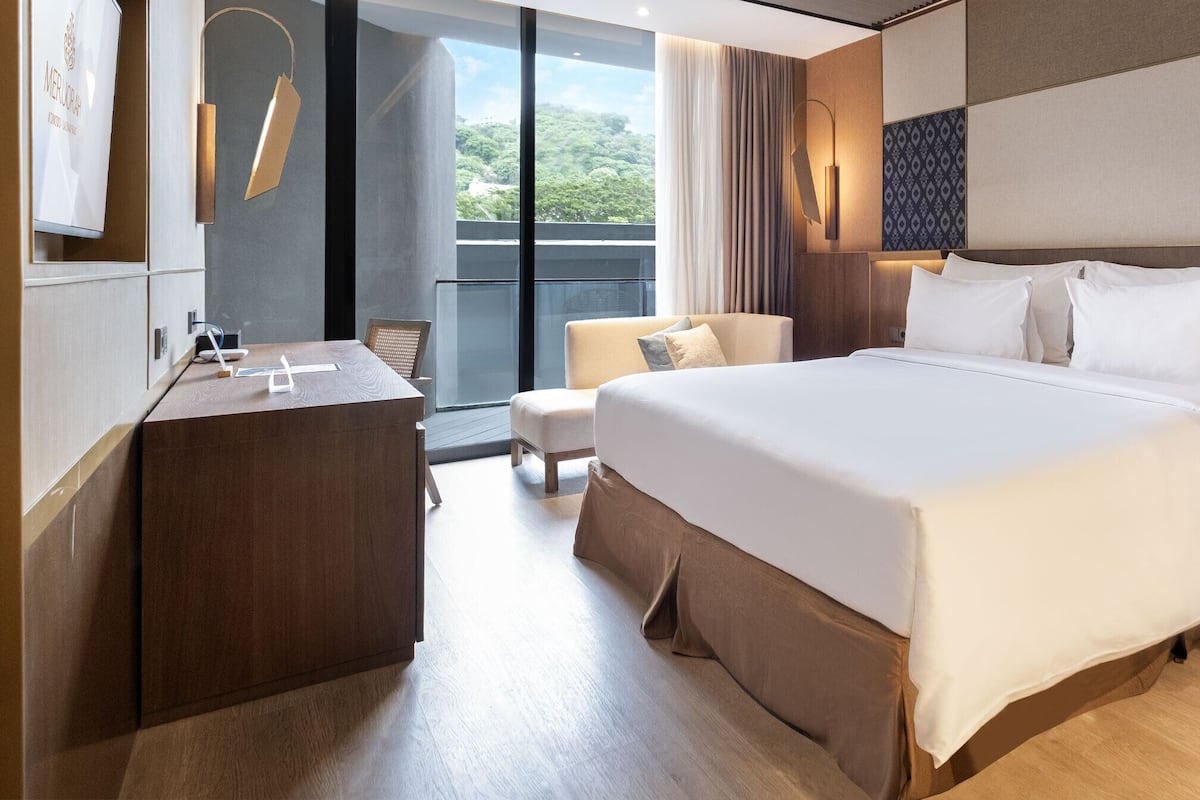 A modern hotel room with a large bed, a desk, and a view of greenery outside.
