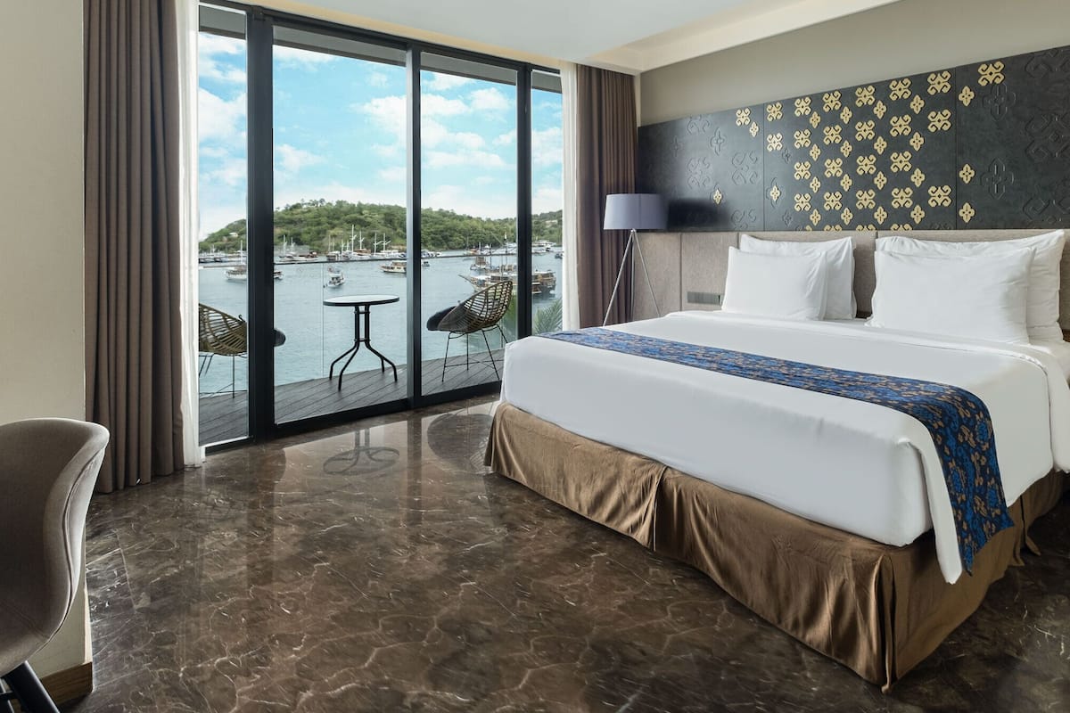 A hotel room with a large bed, a view of the marina, a chair, a table, and a nightstand with a lamp.