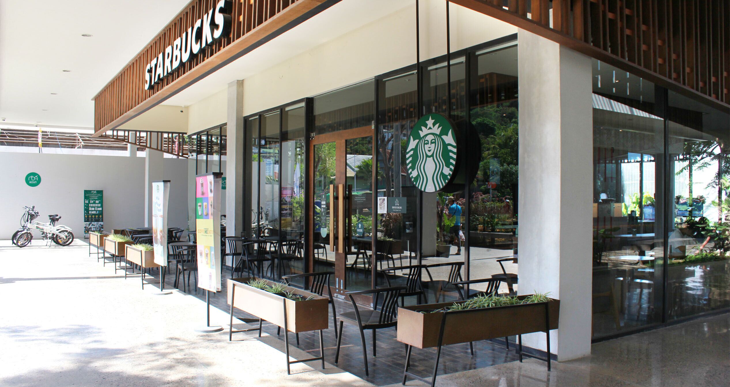 A Starbucks store with outdoor seating, a bike parked outside, and a sign indicating a nearby restaurant.