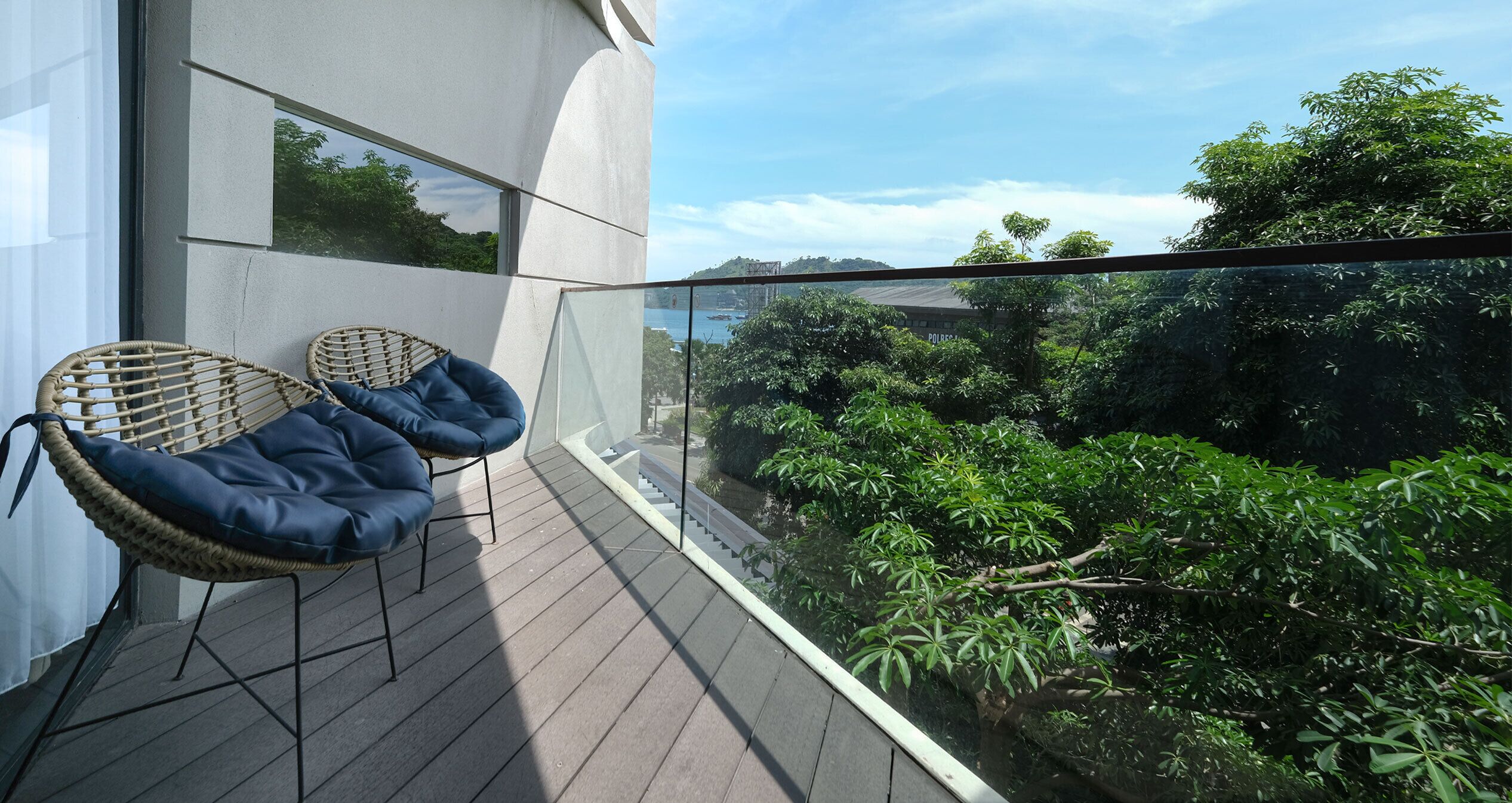 Un balcón con sillas de mimbre y barandilla de vidrio, con vistas a un jardín exuberante y una vista distante de un puente.