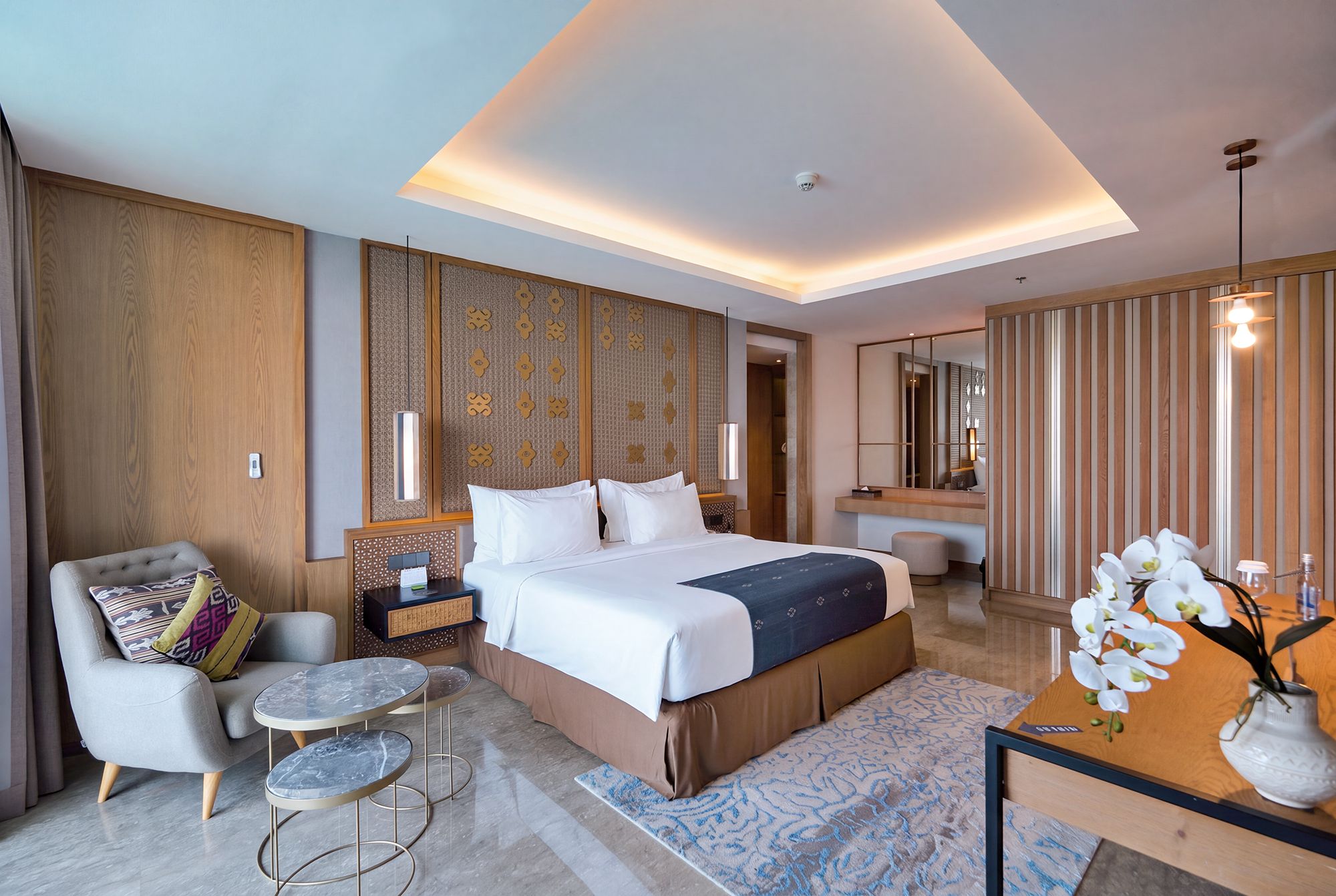 A modern hotel room with a large bed, a sitting area with a chair and a small table, a bathroom with a mirror, and a decorative wall panel.